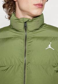 Giacca puffer verde oliva con colletto alto, chiusura con zip e logo Jumpman bianco sul lato sinistro del petto. Texture liscia, design leggero.
