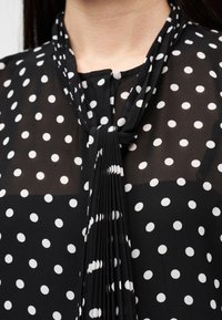 Μαύρη μπλούζα με λευκές πουά, ρεγκλάν λεπτομέρεια στο λαιμό με πιέτες και άνοιγμα-κλειδί στον γιακά. Διάφανο ύφασμα με υφή μοτίβο.