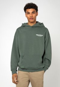 Dunkelgrüne Kapuzenjacke aus weichem Material mit einer vorderen Tasche, gerippten Bündchen und dem Schriftzug "URBAN EDITION" auf der Brust.