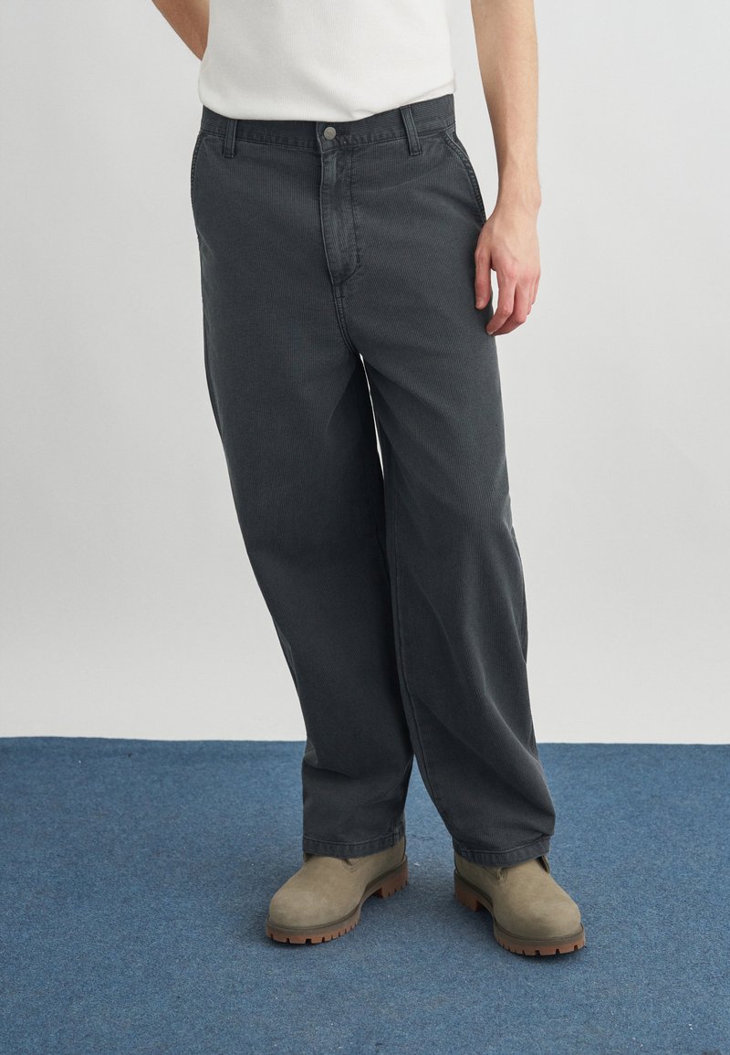 Uomo che indossa pantaloni larghi grigio scuro, stivali da lavoro beige e una canottiera bianca, in piedi su un tappeto blu con sfondo grigio.