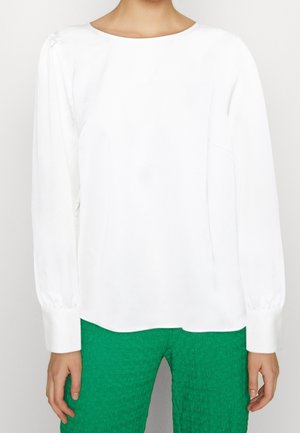 Kobieta w białej bluzce z długimi rękawami z bufkami na mankietach i zielonych teksturowanych spodniach, stoi z ramionami swobodnie opuszczonymi wzdłuż ciała.