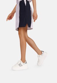 Sneakers bianchi con suola spessa, caratterizzati da dettagli argentati sul tallone e sul lato. Texture liscia con lacci e dettagli di design sottili.