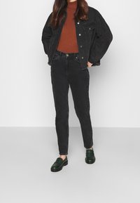 Osoba oblečená v černé džínové bundě, měděném roláku, černých kalhotách s vysokým pasem a tmavozelených mokasínech stojí na bílém pozadí.