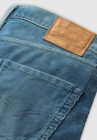 Material din corduroy bleu deschis, cu un model de nervuri vizibil, având o etichetă din piele maro cu branding embosat și detalii de cusătură subtile.