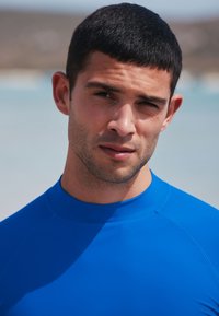 Blå rashguard lavet af elastisk stof med høj hals og glat tekstur. Baggrunden viser en lys strandscene.