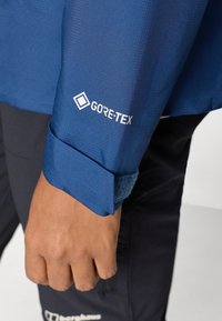 Modrá GORE-TEX bunda s textúrovanou látkou na rukáve, so priliehavým manžetovým zakončením a nastaviteľným suchým zipsom. Tmavé nohavice viditeľné nižšie.