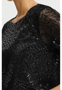 Haut noir en tissu texturé avec des motifs de sequins, col rond et manches transparentes. Le design présente un motif abstrait en ondulation.