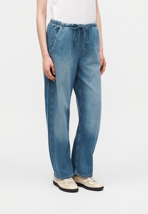 Persoană care poartă pantaloni largi din denim albastru deschis cu talie elastică și șnur, top alb și adidași bej pe un fundal simplu.