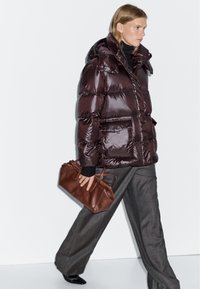 Brown puffer jacket with a shiny surface, large hood, front pockets, and high collar, paired with brown leather bag and gray trousers.