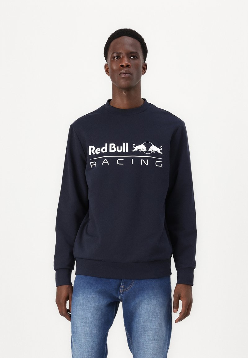 Tamno plava dukserica izrađena od pamuka, s bijelim logotipom "Red Bull Racing" preko prsnog dijela, okruglim izrezom i rebrastim manžetama.