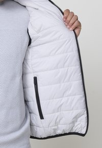 Weiße Daunenjacke mit gestepptem Muster, Innentasche, schwarzem Reißverschluss und kontrastierendem grauem Innenfutter. Glatte Oberfläche und leichtes Design.