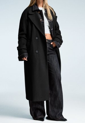 Femme portant un long manteau noir par-dessus une veste en jean noire, un top court blanc, un jean noir à jambes larges et des chaussures noires pointues.
