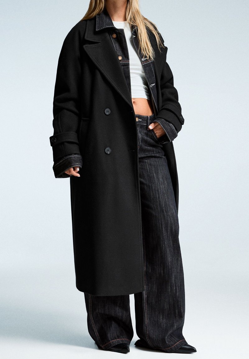 Femme portant un long manteau noir par-dessus une veste en jean noire, un top court blanc, un jean noir à jambes larges et des chaussures noires pointues.