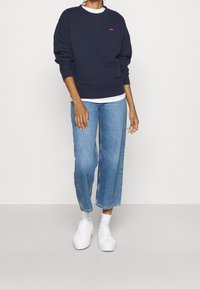 Námořnická mikina s kulatým výstřihem a logem, zkombinovaná s širokými modrými džíny a bílými teniskami, prezentující ležérní a uvolněný styl.