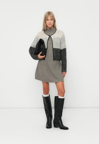 Γυναίκα που φοράει ένα γκρι φόρεμα με ψηλό λαιμό, πολύχρωμο cardigan, μαύρες μπότες μέχρι το γόνατο, κρατώντας μια μαύρη τσάντα clutch, στέκεται μπροστά σε έναν λευκό τοίχο.