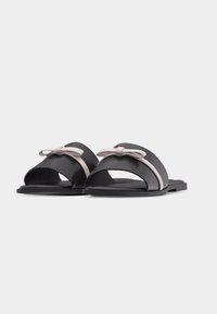 Paire de sandales plates noires à larges brides avec petits nœuds rose clair sur le dessus sur un fond blanc uni.