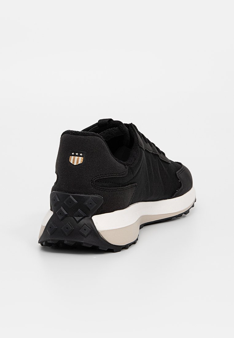 Zapatilla negra con una parte superior de tejido texturizado, diseño de cordones, entresuela blanca en contraste y suela de goma con patrón. Incluye un parche con el logo.