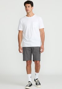 White short-sleeve t-shirt with a chest pocket paired with gray shorts. Model wears white socks and dark sneakers. Simple design, casual fit.