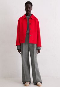 Rode jas van wolmix met grote zakken, grijze gebreide trui en grijze wijde broek. Crèmekleurige sneakers met rode accenten maken de look compleet.