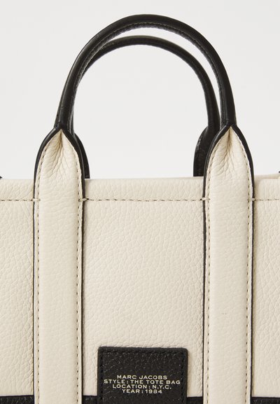 White leather tote bag with black accents, structured shape, short handles, and a black label detailing brand and style information.