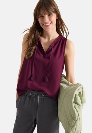 Uśmiechnięta kobieta w bezrękawnikowej bordowej bluzce i ciemnych spodniach, trzymająca jasnobeżową pikowaną kurtkę na jednym ramieniu na jednolitym tle.