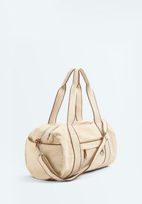 Béžová plátenná taška typu duffel s hnedým lemovaním, dvojitými rukoväťami, nastaviteľným ramenným popruhom a zipsovými bočnými vreckami na bielom pozadí.