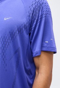 Κοντινή λήψη ενός ατόμου που φοράει μοβ αθλητική μπλούζα Nike με μικρές οπές και μαύρα γραφικά σχέδια στο ύφασμα.