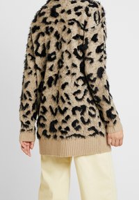 Flauschiger, pelzähnlicher Pullover in Beige mit schwarzem Leopardenmuster, der einen gerippten Saum und Ärmelsäume aufweist. Das Design auf der Rückseite betont das Tiermuster.