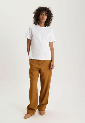 Femme aux cheveux bouclés debout, portant un t-shirt blanc à manches courtes, un pantalon marron ample et des chaussures plates beiges sur un fond uni.