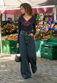 Vrouw met krullend haar, gekleed in een zwarte langemouwenshirt en wijde jeans, met een zwarte ronde handtas, die langs een fruitkraam buiten wandelt.