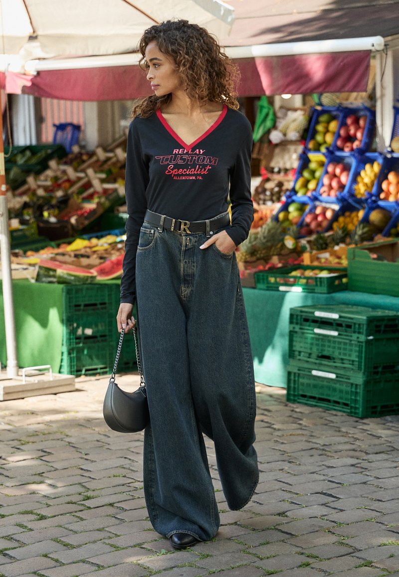 Vrouw met krullend haar, gekleed in een zwarte langemouwenshirt en wijde jeans, met een zwarte ronde handtas, die langs een fruitkraam buiten wandelt.