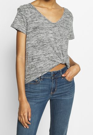 Kvinde iført en grå marmoreret kortærmet skjorte, bundet i taljen, kombineret med blå denimjeans, hånd i lommen.