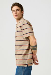 Beige kortärmad polo skjorta med horisontella röda och marinblå ränder, med krage och ett litet emblem på vänster bröst, samt sidofack.