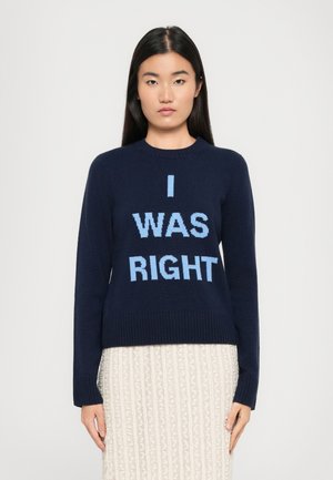 Une femme aux longs cheveux noirs porte un pull marine avec "J'AVAIS RAISON" en lettres bleu clair et une jupe crème texturée, se tenant contre un fond blanc.
