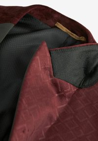 Interiorul unei jachete burgundy, realizat dintr-un material texturat, cu o captuseala neagra si un model geometric subtil. Finisaj neted si forma croita vizibile.