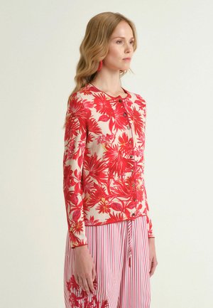 Donna con lunghi capelli biondi che indossa un cardigan a fiori rosso e crema con bottoni e pantaloni a righe rosse e bianche con ricami floreali.
