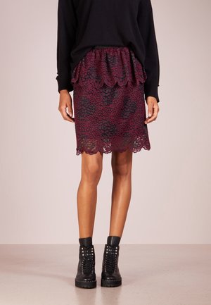 Femme portant un pull noir, une jupe en dentelle bordeaux avec des volants, et des bottines noires à lacets, debout sur un sol neutre.