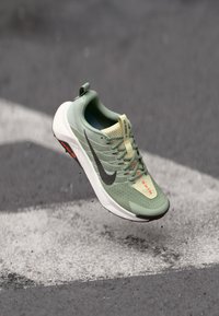 Svetlozelené bežecké topánky s textilným zvrškom, sivý logo Nike a biela podrážka, so zvýrazneniami a šnúrkami v oranžovej farbe. Ľahký dizajn pre optimálny výkon.