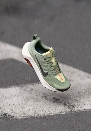 Lys grønt løbesko med mesh-overdel, grå Nike-svob, og hvid sål, med orange accenter og snørebånd. Letvægtsdesign til præstation.