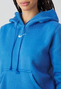 Närbild av en person som bär en ljusblå Nike-hoodie med dragsnören och vit swoosh-logga centrerad på bröstet, händerna i framfickan.