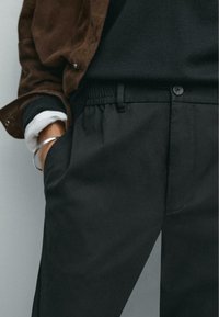 Zwarte broek met een elastische tailleband, een knoop aan de voorkant, zijzakken en een gladde textuur, gecombineerd met een bruine suède jasje.