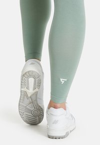 Light green leggings with a smooth texture, featuring a white logo on the calf, paired with white athletic shoes with gray soles.