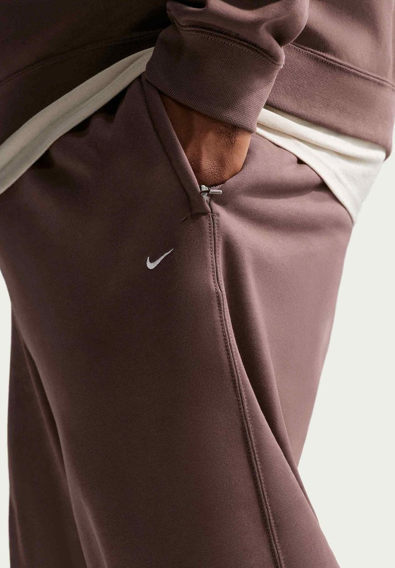 Persoon draagt mauve Nike-joggingbroek en sweatshirt met hand in de zak, wit shirt zichtbaar onder de sweatshirt. Nike-logo op de broek.