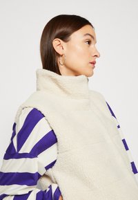 Fleece pullover in crème met een hoge kraag, voorzien van een textuur stof en gelaagd ontwerp. Draag het over een paars met witte gestreepte top.
