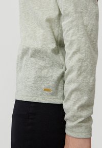 Light green long-sleeve shirt made from textured fabric, featuring a small gold logo tag near the hem. Black pants visible below.