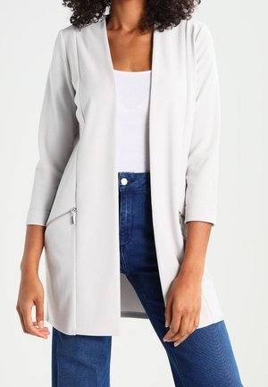 Kvinde iført en lysegrå åben blazer med lynlåslommer over en hvid top og blå jeans mod en ensfarvet baggrund.