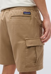 Μπεζ καμβά σορτς με πίσω και πλαϊνές τσέπες με καπάκι, λογότυπο Patagonia πάνω από την πίσω τσέπη, φορεμένα από ένα άτομο με το χέρι κοντά στην πλαϊνή τσέπη.