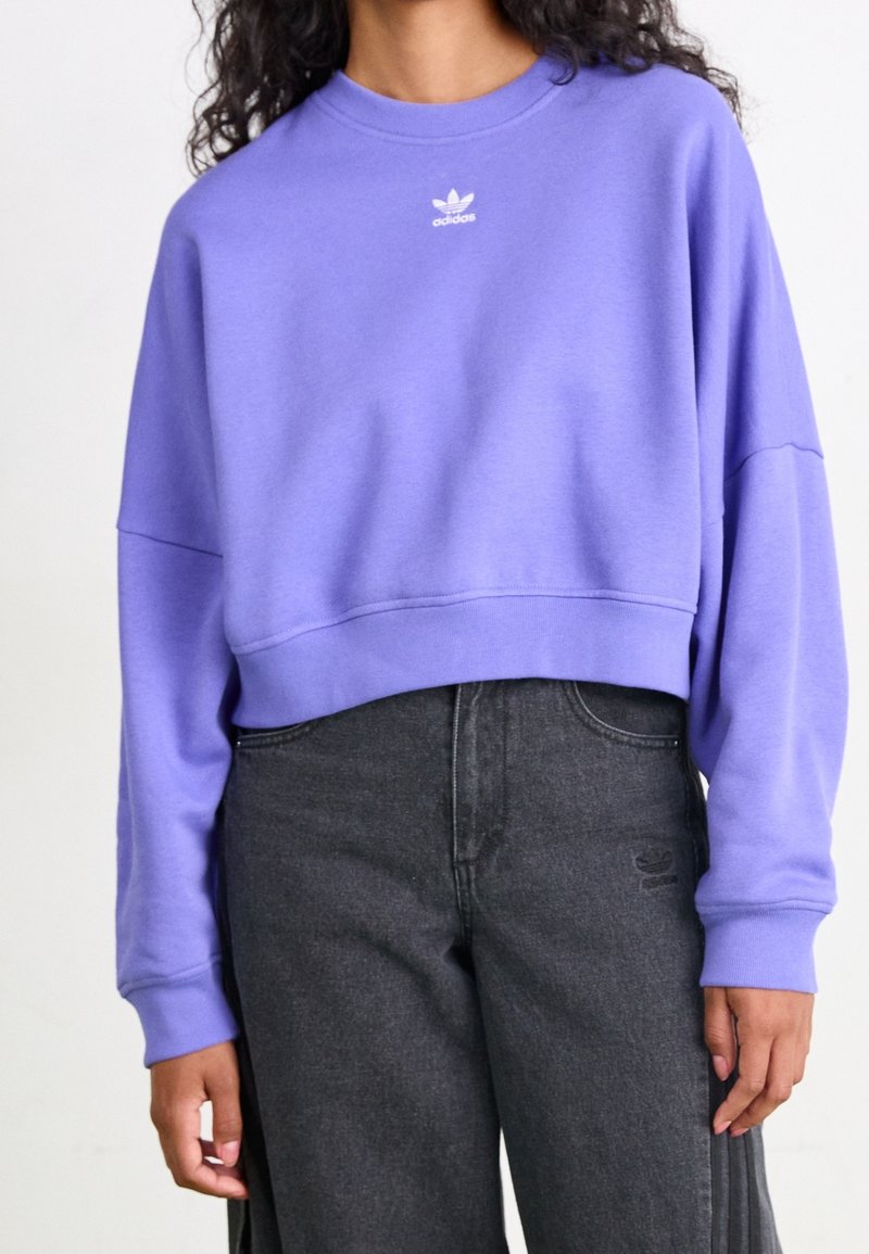Sudadera corta púrpura con cuello redondo, puños acanalados y pequeño logo blanco de Adidas. Combinada con jeans oscuros de tiro alto.