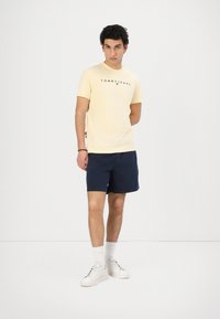 Gelbes Kurzarm-T-Shirt mit "TOMMY JEANS"-Logo, kombiniert mit marineblauen Shorts, weißen Socken und Sneakers. Das Modell posiert vor einem schlichten Hintergrund.