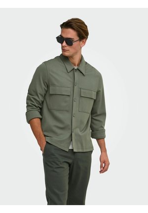 Homme portant une chemise à boutons vert foncé avec deux poches poitrine et des lunettes de soleil foncées, debout avec une main dans la poche.